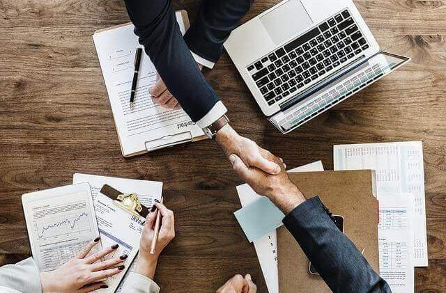 Two business people shake hands, while a third observes. This happens over a wooden table covered in various paperwork, and a laptop is sat to the right of one of the businessfolk.
