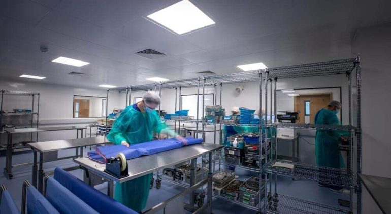 Workers busy sterlising medical tools in a storeroom, lined with sanitary surface covers over steel tables, and metal racks.