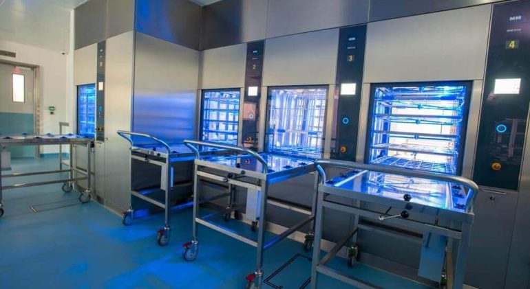 A surgical equipment sterilisation centre, with empty trolleys sitting against UV sterilisation machines. The floor is blue, the machines are steel silver, and there is a white and blue door in the background.
