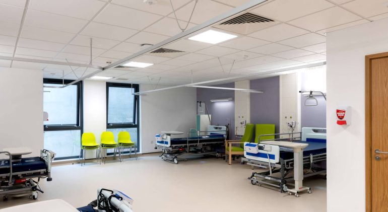 Inside a recovery ward at a hospital, with purple walls and clean beds, sidetables, chairs and lights about.