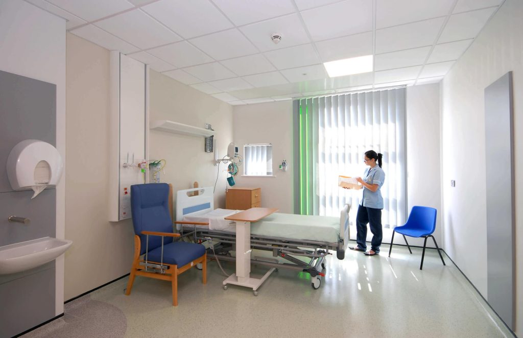 The inside of a ward, with a clean bed, side table, chair, and a healthcare helper present.