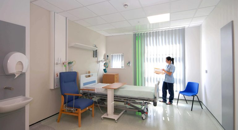 The inside of a ward, with a clean bed, side table, chair, and a healthcare helper present.